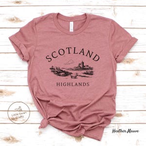 May include: Mauve t-shirt featuring a black illustration of a Scottish scene with a castle, sheep, and the text "SCOTLAND HIGHLANDS." The shirt has a crew neck and short sleeves. The brand name "Little Swan Crafts" is visible.