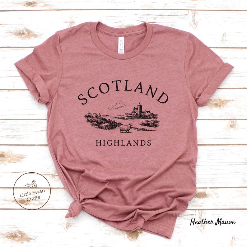 May include: Mauve t-shirt featuring a black illustration of a Scottish scene with a castle, sheep, and the text "SCOTLAND HIGHLANDS." The shirt has a crew neck and short sleeves. The brand name "Little Swan Crafts" is visible.