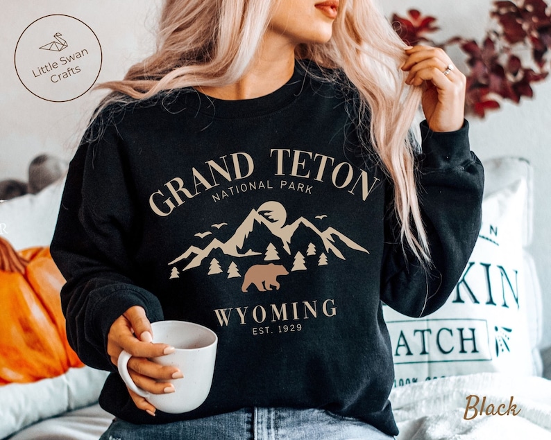 May include: Black sweatshirt with a tan graphic design featuring the text "GRAND TETON NATIONAL PARK" and a mountain scene with a bear. The text "WYOMING EST. 1929" is below the graphic.