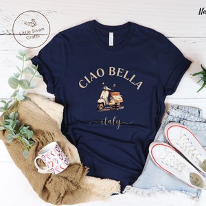 Pu&ograve; includere: Una maglietta blu navy con il testo "Ciao Bella" in un font color sabbia, un'immagine a cartone animato di uno scooter bianco e la parola "italy" in un font corsivo color sabbia.