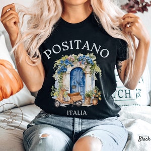 Puede incluir: Camiseta negra con un gr&aacute;fico blanco de una puerta azul con flores y un scooter delante. El texto "Positano Italia" est&aacute; impreso debajo del gr&aacute;fico.