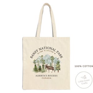 May include: A beige canvas tote bag featuring a scenic illustration of Banff National Park. The design includes mountains, trees, and a moose, with text that reads "BANFF NATIONAL PARK, ALBERTA'S ROCKIES, CANADA". The bag is made of 100% cotton.