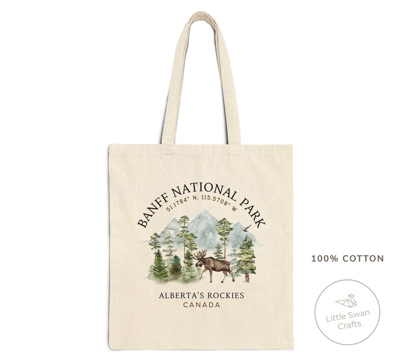 May include: A beige canvas tote bag featuring a scenic illustration of Banff National Park. The design includes mountains, trees, and a moose, with text that reads "BANFF NATIONAL PARK, ALBERTA'S ROCKIES, CANADA". The bag is made of 100% cotton.