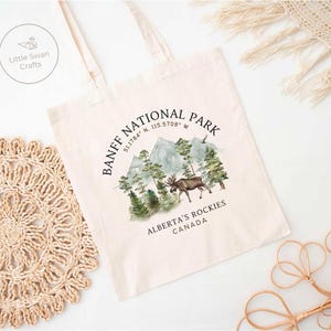 May include: Cream-colored tote bag featuring a watercolor design of Banff National Park. The design includes mountains, trees, and a moose, with text that reads "BANFF NATIONAL PARK, ALBERTA'S ROCKIES, CANADA". The bag has long handles.