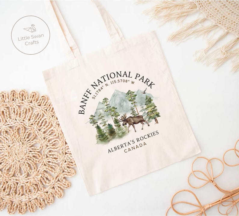 May include: Cream-colored tote bag featuring a watercolor design of Banff National Park. The design includes mountains, trees, and a moose, with text that reads "BANFF NATIONAL PARK, ALBERTA'S ROCKIES, CANADA". The bag has long handles.