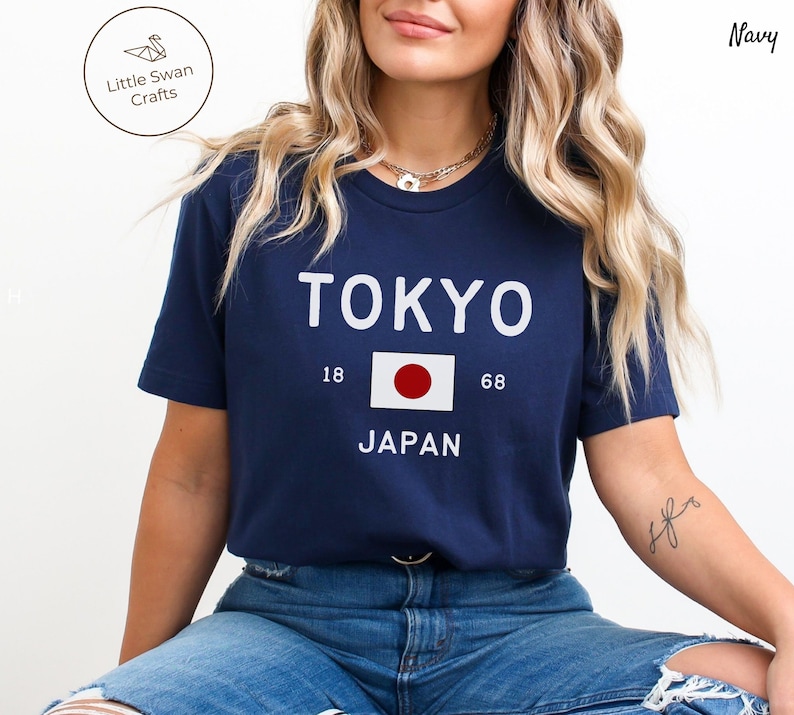 K&ouml;nnte beinhalten: Ein marineblaues T-Shirt mit wei&szlig;em Text, der "TOKYO" und "JAPAN" lautet, mit der japanischen Flagge dazwischen. Die Zahlen 18 und 68 befinden sich auf beiden Seiten der Flagge.