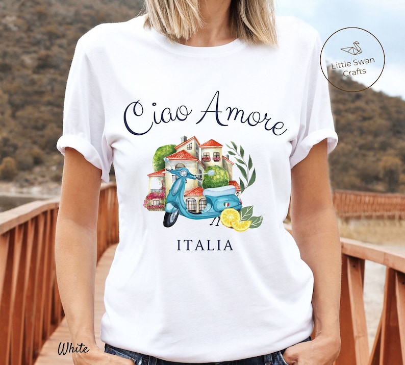 May include: White t-shirt with a blue and white scooter, a small building with red roofs, and green plants. The text on the shirt reads 'Ciao Amore' and 'ITALIA'.