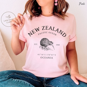 Peut inclure: T-shirt rose avec un motif graphique noir repr&eacute;sentant un kiwi, le texte "NEW ZEALAND", "PACIFIC OCEAN", "EST. 1840", "41&deg;18&prime;S 174&deg;47&prime;E", et "OCEANIA".