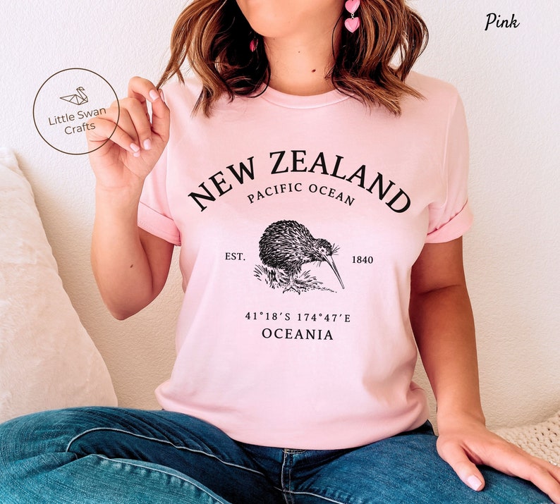 Peut inclure: T-shirt rose avec un motif graphique noir repr&eacute;sentant un kiwi, le texte "NEW ZEALAND", "PACIFIC OCEAN", "EST. 1840", "41&deg;18&prime;S 174&deg;47&prime;E", et "OCEANIA".