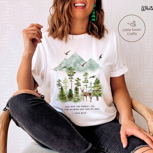 Pode incluir: Camiseta branca com uma ilustra&ccedil;&atilde;o em aquarela de uma cena de floresta com montanhas, &aacute;rvores e um veado. O texto "AND INTO THE FOREST I GO. TO LOSE MY MIND AND FIND MY SOUL - JOHN MUIR -" est&aacute; impresso na camiseta.