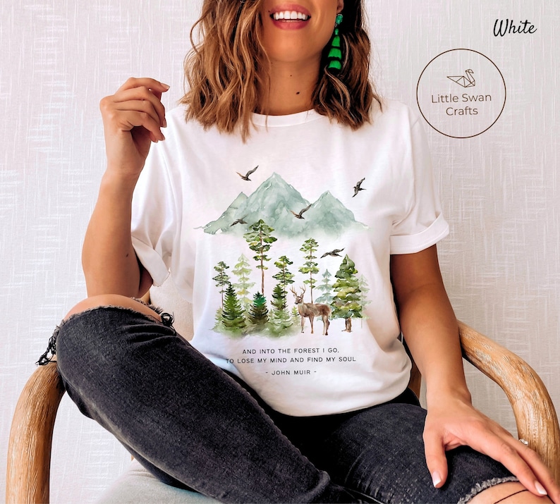 Pode incluir: Camiseta branca com uma ilustra&ccedil;&atilde;o em aquarela de uma cena de floresta com montanhas, &aacute;rvores e um veado. O texto "AND INTO THE FOREST I GO. TO LOSE MY MIND AND FIND MY SOUL - JOHN MUIR -" est&aacute; impresso na camiseta.