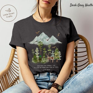 Pode incluir: Camiseta em cinza escuro mesclado com uma ilustra&ccedil;&atilde;o em aquarela de uma cordilheira, uma floresta e um veado. O texto "AND INTO THE FOREST I GO TO LOSE MY MIND AND FIND MY SOUL - JOHN MUIR -" est&aacute; impresso na camiseta.