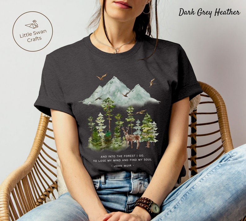 Pode incluir: Camiseta em cinza escuro mesclado com uma ilustra&ccedil;&atilde;o em aquarela de uma cordilheira, uma floresta e um veado. O texto "AND INTO THE FOREST I GO TO LOSE MY MIND AND FIND MY SOUL - JOHN MUIR -" est&aacute; impresso na camiseta.