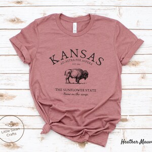 May include: A heather mauve t-shirt with a black graphic design featuring the text "KANSAS", "AD ASTRA PER ASPERA", "EST. 1861", "THE SUNFLOWER STATE", and "Home on the range". The graphic also includes a black and white illustration of a buffalo.