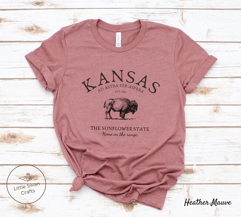 May include: A heather mauve t-shirt with a black graphic design featuring the text "KANSAS", "AD ASTRA PER ASPERA", "EST. 1861", "THE SUNFLOWER STATE", and "Home on the range". The graphic also includes a black and white illustration of a buffalo.