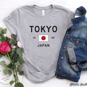 K&ouml;nnte beinhalten: Graues T-Shirt mit dem Text "TOKYO" in Schwarz, einem roten Kreis auf wei&szlig;em Hintergrund, den Zahlen "18" und "68" und dem Wort "JAPAN" in Schwarz.