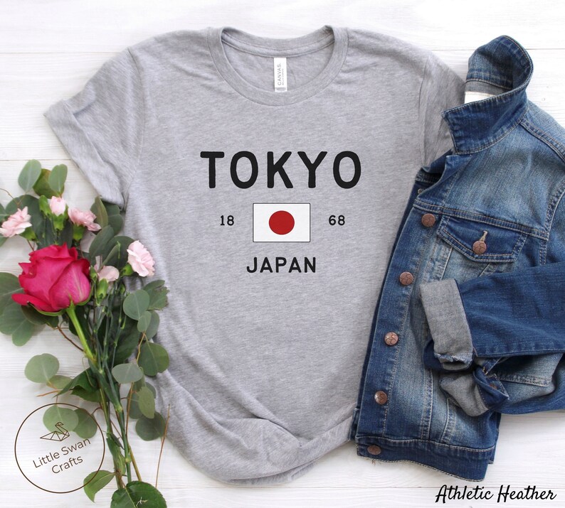 K&ouml;nnte beinhalten: Graues T-Shirt mit dem Text "TOKYO" in Schwarz, einem roten Kreis auf wei&szlig;em Hintergrund, den Zahlen "18" und "68" und dem Wort "JAPAN" in Schwarz.