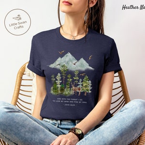 Pode incluir: Uma camiseta azul marinho heather com uma ilustra&ccedil;&atilde;o em aquarela de uma cordilheira, &aacute;rvores e um veado. O texto "AND INTO THE FOREST I GO TO LOSE MY MIND AND FIND MY SOUL - JOHN MUIR -" est&aacute; impresso abaixo da ilustra&ccedil;&atilde;o.