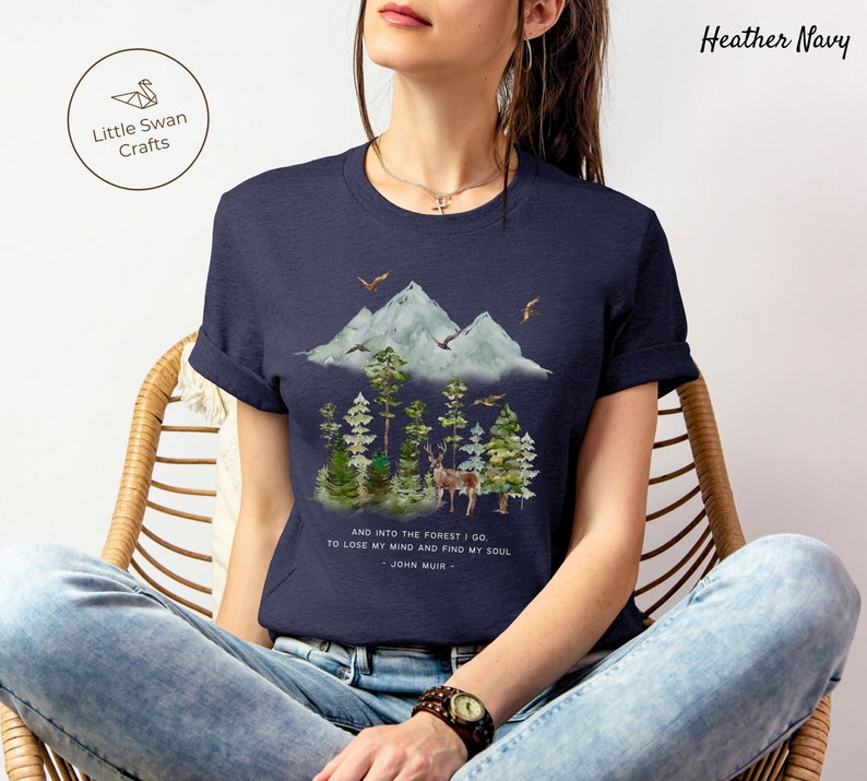 Pode incluir: Uma camiseta azul marinho heather com uma ilustra&ccedil;&atilde;o em aquarela de uma cordilheira, &aacute;rvores e um veado. O texto "AND INTO THE FOREST I GO TO LOSE MY MIND AND FIND MY SOUL - JOHN MUIR -" est&aacute; impresso abaixo da ilustra&ccedil;&atilde;o.