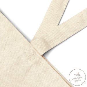 May include: Close-up of a natural canvas tote bag with cream-colored straps. The straps are sewn to the bag's body, forming a V-shape. The bag has a simple, minimalist design, with the "Little Swan Crafts" logo in the lower right corner.