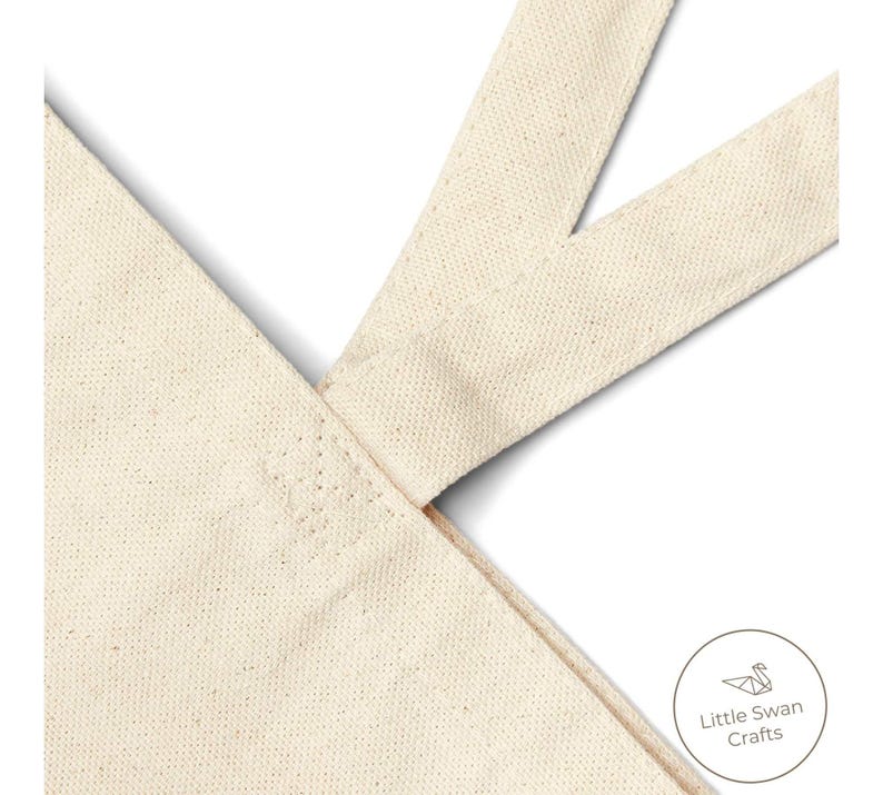May include: Close-up of a natural canvas tote bag with cream-colored straps. The straps are sewn to the bag's body, forming a V-shape. The bag has a simple, minimalist design, with the "Little Swan Crafts" logo in the lower right corner.