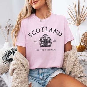 Pode incluir: Camiseta rosa claro com a palavra "SCOTLAND" em preto, arqueada na parte superior. Abaixo, um bras&atilde;o e o texto "EST. 843 UNITED KINGDOM". A camiseta &eacute; feita de um material macio.