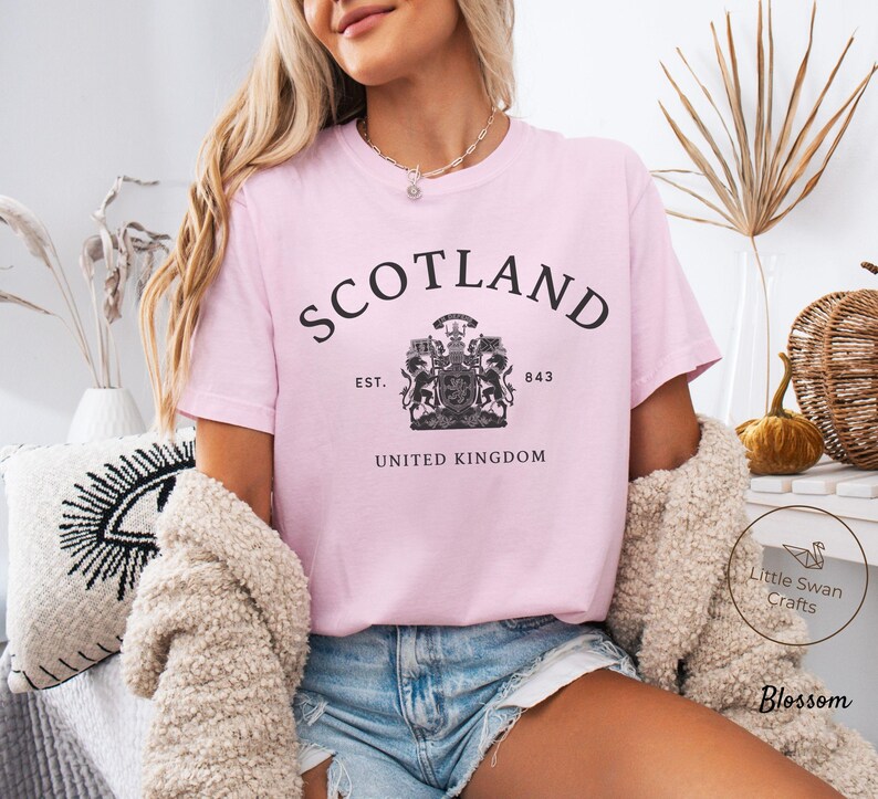 Pode incluir: Camiseta rosa claro com a palavra "SCOTLAND" em preto, arqueada na parte superior. Abaixo, um bras&atilde;o e o texto "EST. 843 UNITED KINGDOM". A camiseta &eacute; feita de um material macio.