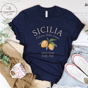 Peut inclure: Un t-shirt bleu marine avec le texte "SICILIA Delicious Italian Lemons FRESH FROM Sicily, Italy" en cr&egrave;me. Un graphique de deux citrons sur une branche est au-dessus du texte.
