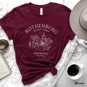 Pode incluir: Camiseta de cor vinho com um design gr&aacute;fico branco da cidade de Rothenburg ob der Tauber na Alemanha. O texto "Rothenburg ob der Tauber", "Alemanha" e "Baviera" est&aacute; impresso em branco na camiseta.