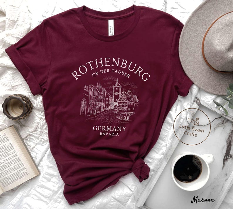 Pode incluir: Camiseta de cor vinho com um design gr&aacute;fico branco da cidade de Rothenburg ob der Tauber na Alemanha. O texto "Rothenburg ob der Tauber", "Alemanha" e "Baviera" est&aacute; impresso em branco na camiseta.