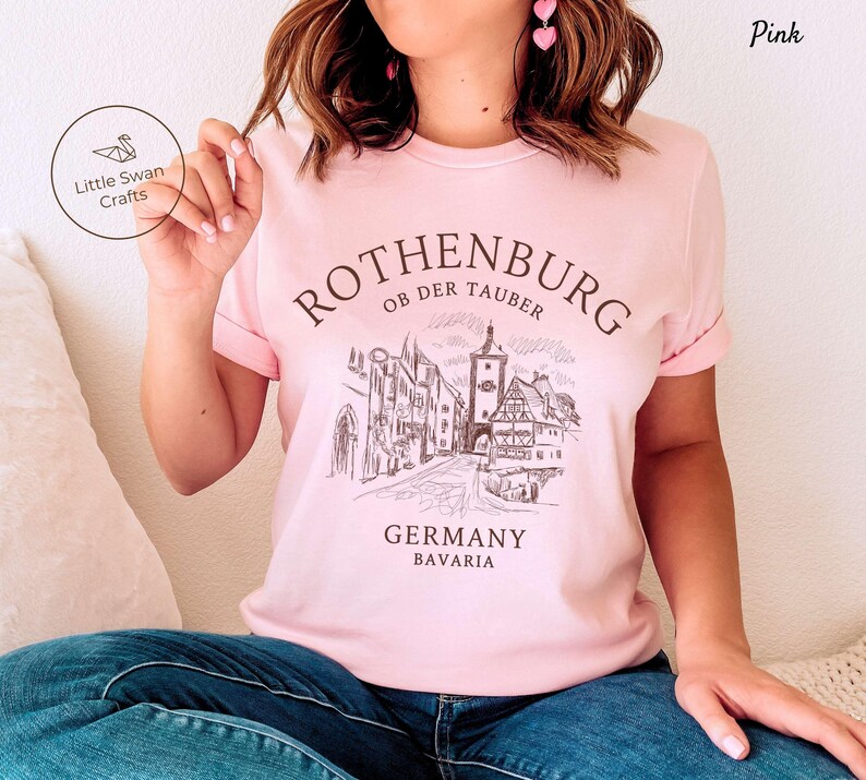 Pode incluir: Camiseta rosa com uma ilustra&ccedil;&atilde;o em preto e branco do horizonte da cidade de Rothenburg ob der Tauber. O texto "Rothenburg ob der Tauber, Alemanha, Baviera" est&aacute; impresso na camiseta.