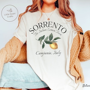 May include: Cream-colored t-shirt with the text "SORRENTO The Italian Lemon Town Campania, Italy" and a lemon illustration. The shirt is part of a clothing collection.