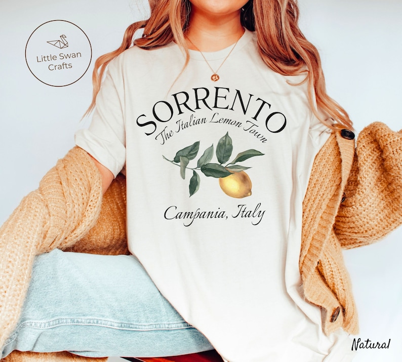 May include: Cream-colored t-shirt with the text "SORRENTO The Italian Lemon Town Campania, Italy" and a lemon illustration. The shirt is part of a clothing collection.