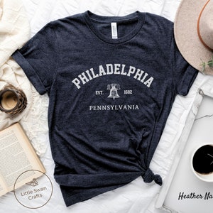 May include: A heather navy blue t-shirt with a white graphic print of the Liberty Bell and the text "Philadelphia, Est. 1682, Pennsylvania".