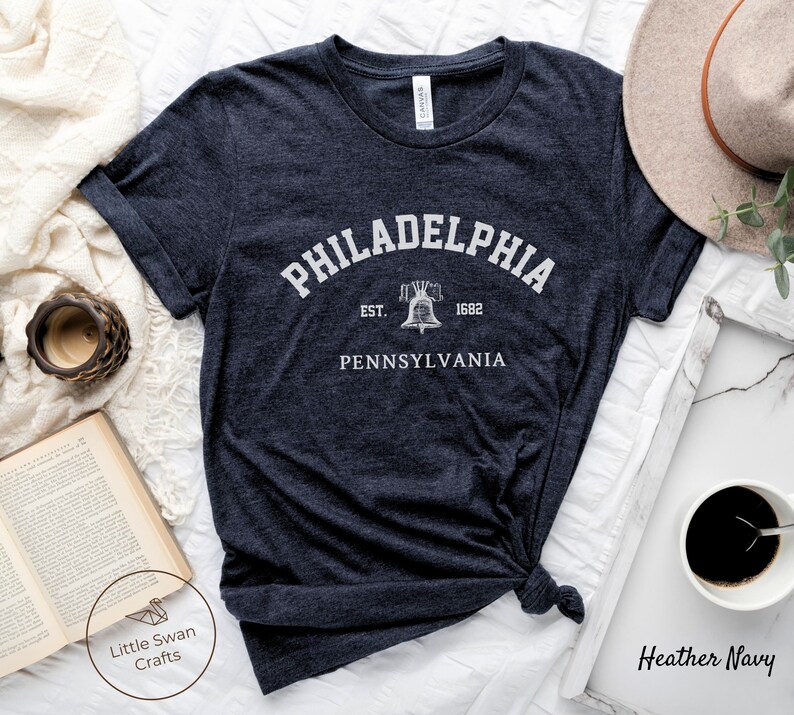 May include: A heather navy blue t-shirt with a white graphic print of the Liberty Bell and the text "Philadelphia, Est. 1682, Pennsylvania".
