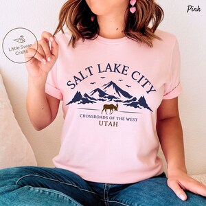 May include: Pink t-shirt with a graphic design of mountains, a moose, and the text "Salt Lake City, Crossroads of the West, Utah".