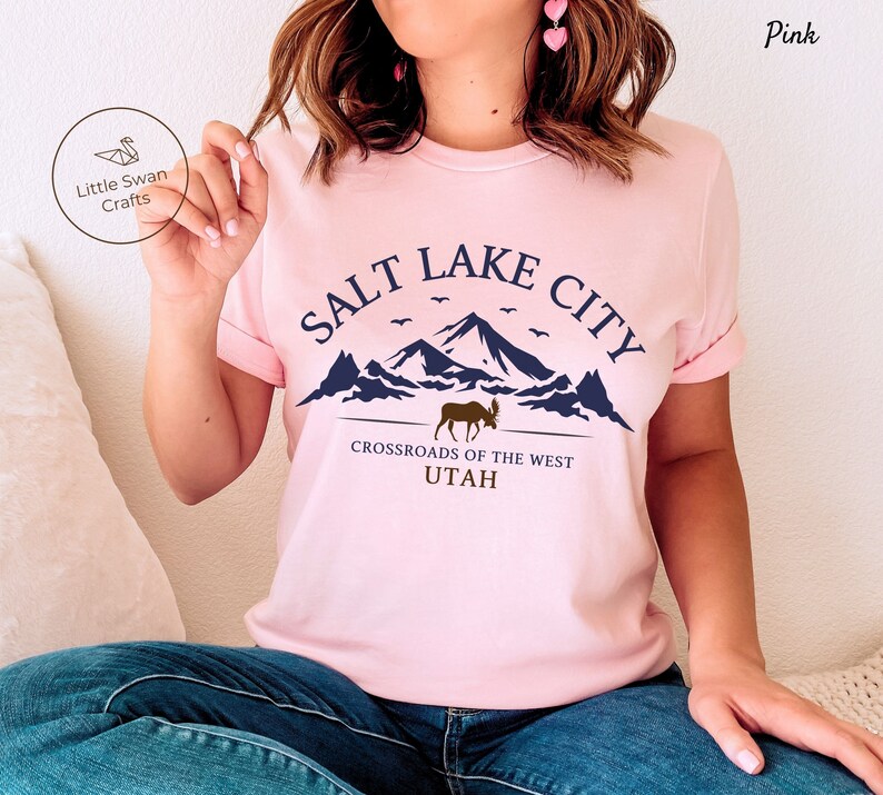 May include: Pink t-shirt with a graphic design of mountains, a moose, and the text "Salt Lake City, Crossroads of the West, Utah".