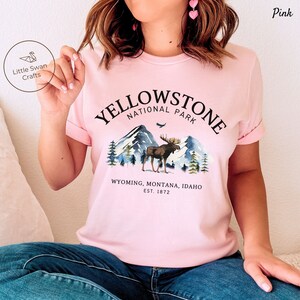 May include: Pink t-shirt featuring a watercolor design of Yellowstone National Park. The graphic includes mountains, a moose, and the text "YELLOWSTONE NATIONAL PARK" and "WYOMING, MONTANA, IDAHO EST. 1872". The brand "Little Swan Crafts" is also visible.