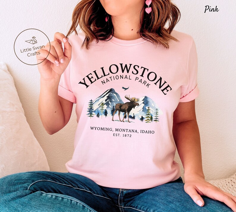 May include: Pink t-shirt featuring a watercolor design of Yellowstone National Park. The graphic includes mountains, a moose, and the text "YELLOWSTONE NATIONAL PARK" and "WYOMING, MONTANA, IDAHO EST. 1872". The brand "Little Swan Crafts" is also visible.