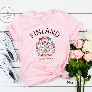 May include: Pink t-shirt with a white graphic design featuring the word "FINLAND" in black text, a circle of stylized flowers, animals, and the word "suomi" in black text.