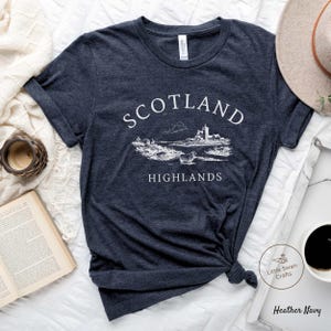May include: Dark navy t-shirt featuring the words "Scotland Highlands" in white, above a detailed landscape illustration. The illustration includes a castle, sheep, and hills. The t-shirt is knotted at the bottom.