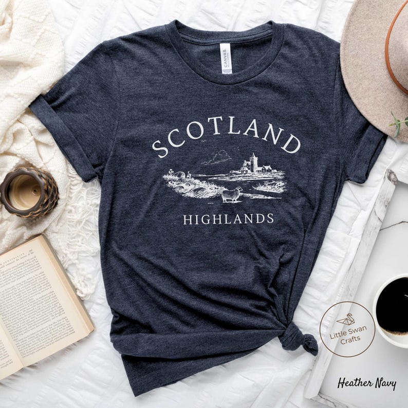 May include: Dark navy t-shirt featuring the words "Scotland Highlands" in white, above a detailed landscape illustration. The illustration includes a castle, sheep, and hills. The t-shirt is knotted at the bottom.