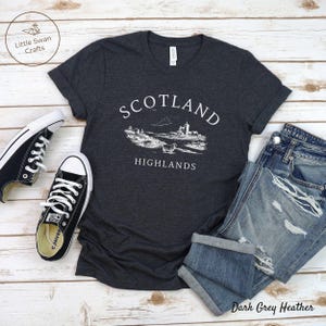 May include: Dark grey heather t-shirt featuring a white graphic of the Scottish Highlands, with the words "Scotland Highlands." The image includes blue jeans and black trainers.