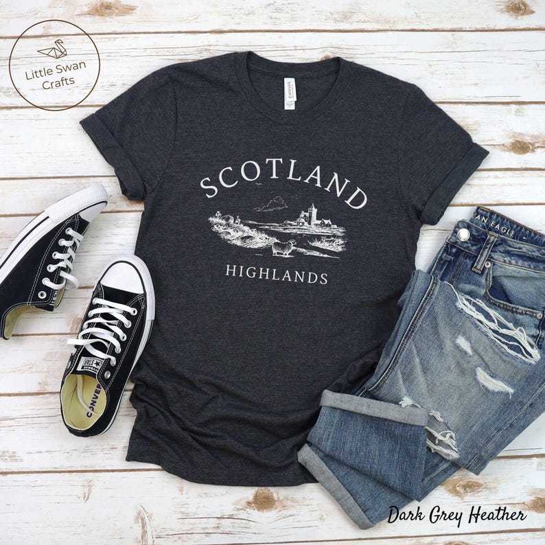 May include: Dark grey heather t-shirt featuring a white graphic of the Scottish Highlands, with the words "Scotland Highlands." The image includes blue jeans and black trainers.