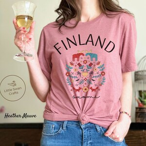 May include: A heather mauve t-shirt with a graphic design featuring the word "FINLAND" in black text, a floral design with moose, foxes, and the word "suomi" in black text.