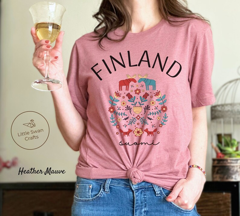 May include: A heather mauve t-shirt with a graphic design featuring the word "FINLAND" in black text, a floral design with moose, foxes, and the word "suomi" in black text.