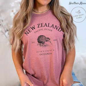 Peut inclure: Un t-shirt en mauve chin&eacute; avec un motif graphique noir repr&eacute;sentant un kiwi et le texte "NEW ZEALAND, EST. 1840, PACIFIC OCEAN, 41&deg;18&prime;S 174&deg;47&prime;E, OCEANIA".