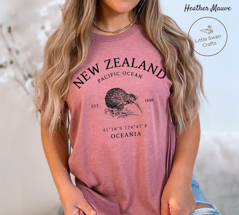 Peut inclure: Un t-shirt en mauve chin&eacute; avec un motif graphique noir repr&eacute;sentant un kiwi et le texte "NEW ZEALAND, EST. 1840, PACIFIC OCEAN, 41&deg;18&prime;S 174&deg;47&prime;E, OCEANIA".