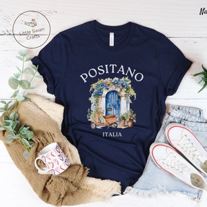 Puede incluir: Camiseta azul marino con un gr&aacute;fico blanco de una puerta azul con flores y un scooter. El texto "Positano Italia" est&aacute; impreso debajo del gr&aacute;fico.