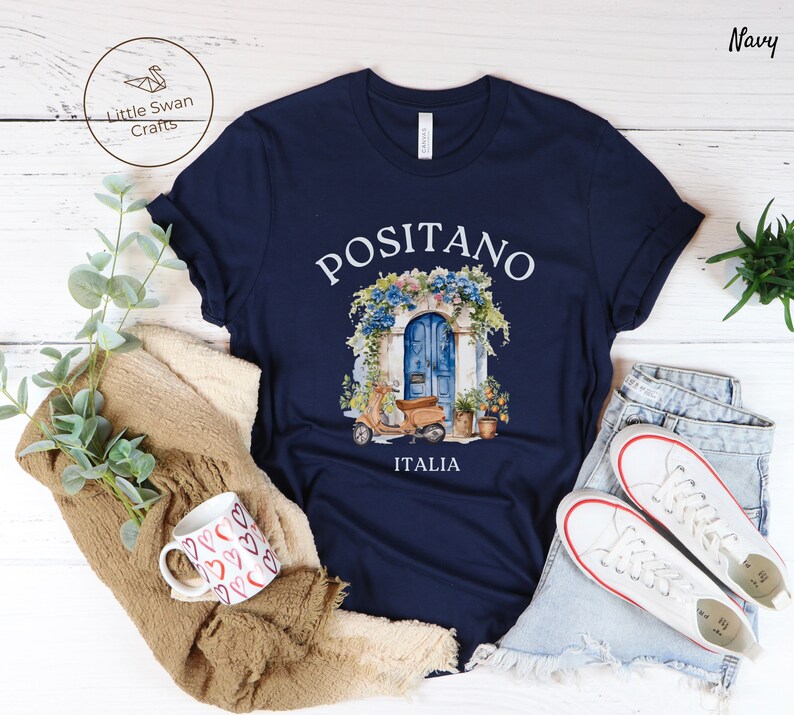 Puede incluir: Camiseta azul marino con un gr&aacute;fico blanco de una puerta azul con flores y un scooter. El texto "Positano Italia" est&aacute; impreso debajo del gr&aacute;fico.